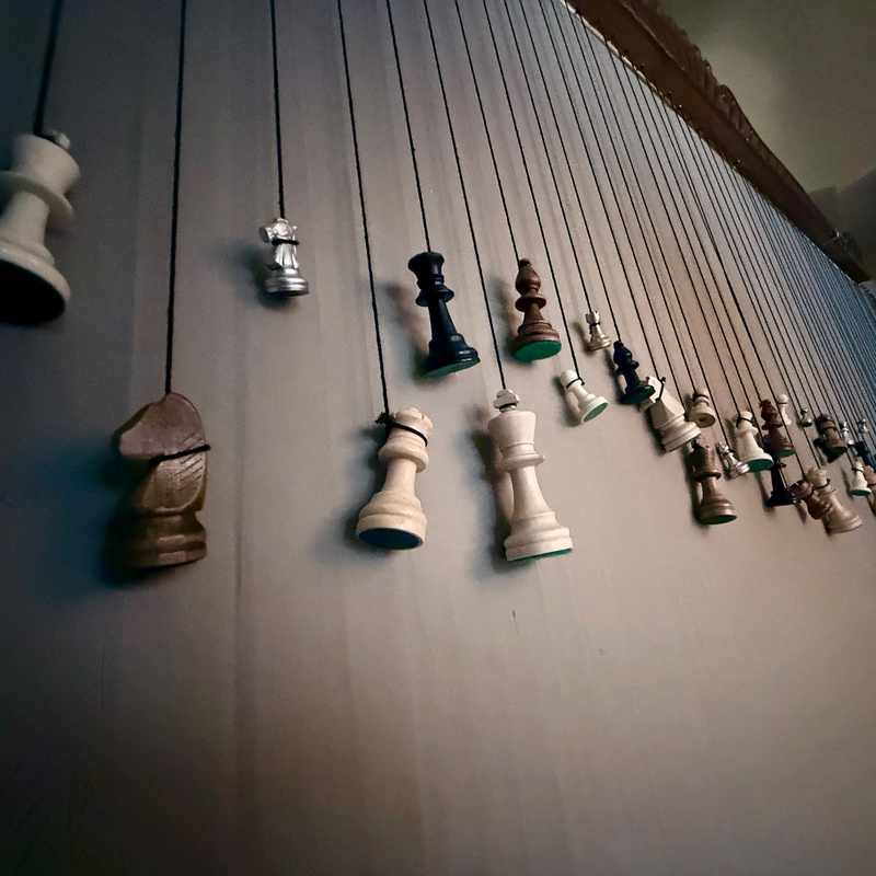 Chess pieces are suspended in a diagonal line, hanging from strings against a wall.