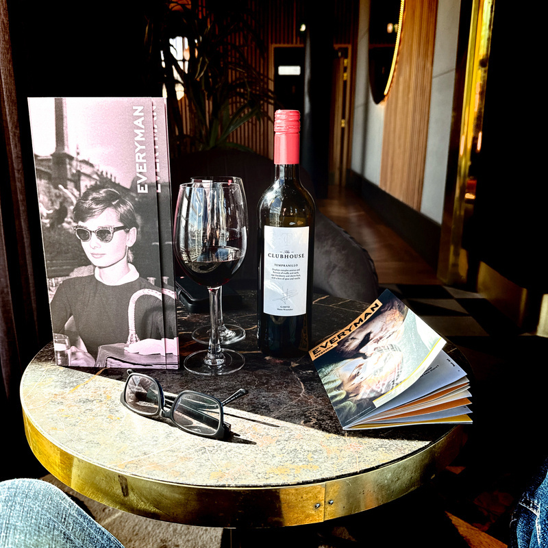 Auto-generated description: A cozy setting features a round table with a wine bottle, a glass of red wine, a book, a pair of glasses, and a magazine with a stylish cover.