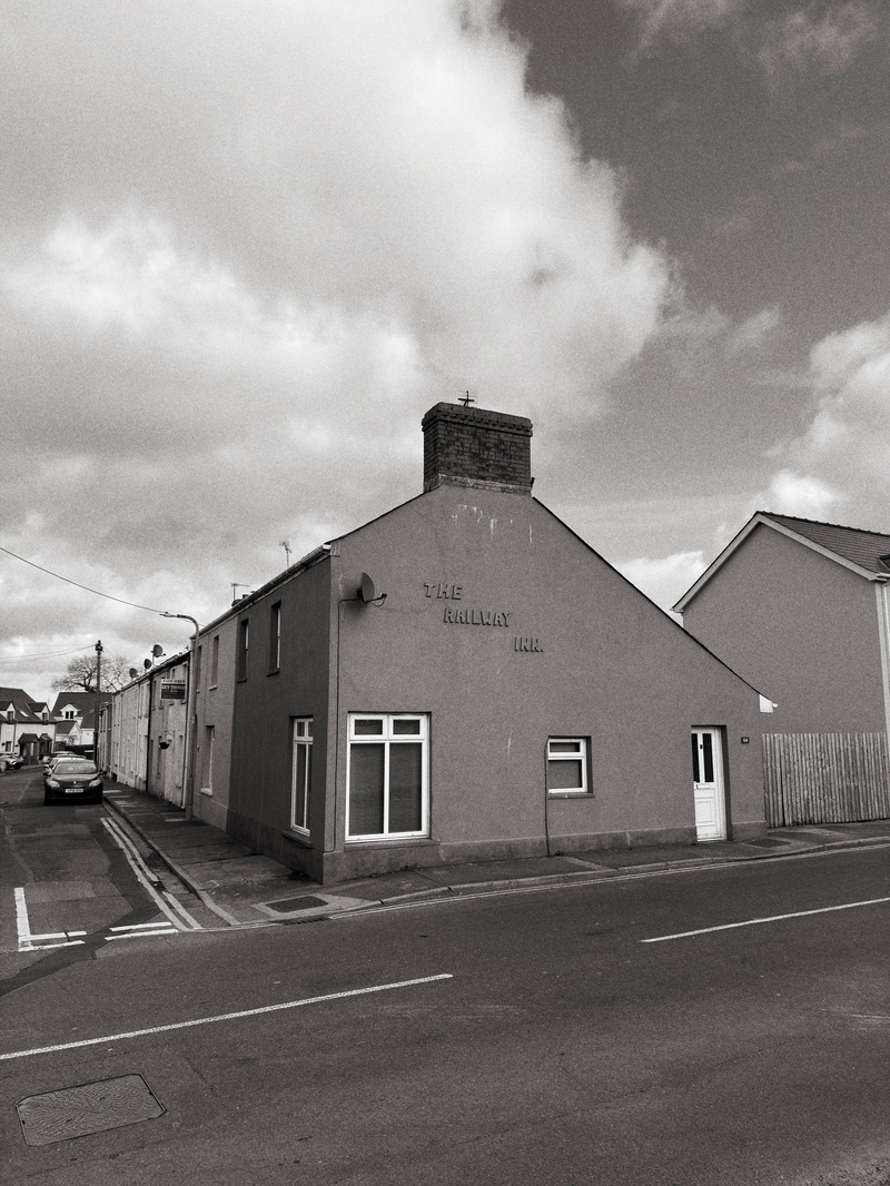 Auto-generated description: A road runs alongside a building marked The Railway Inn, with several other structures and parked cars in the background under a cloudy sky.