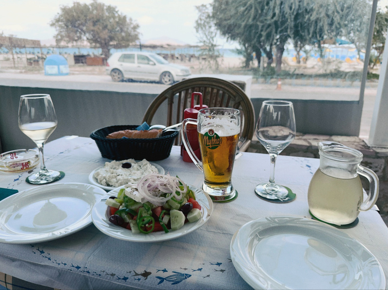 A table is set with plates, glasses of wine and beer, a salad, bread, and a carafe, with a scenic view of trees, cars, and a body of water in the background.
