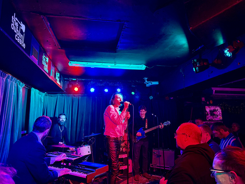Auto-generated description: A live band is performing on stage in a dimly lit venue with colorful lights illuminating the scene.