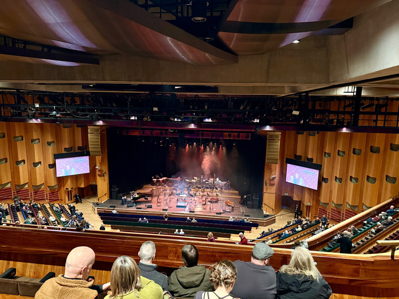 Auto-generated description: An audience sits in a concert hall with a stage set up for a performance, featuring lighting and musical instruments.