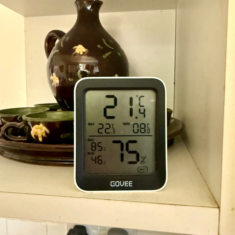 Auto-generated description: A digital thermometer displaying temperature and humidity is placed on a shelf next to ceramic teacups and saucers.