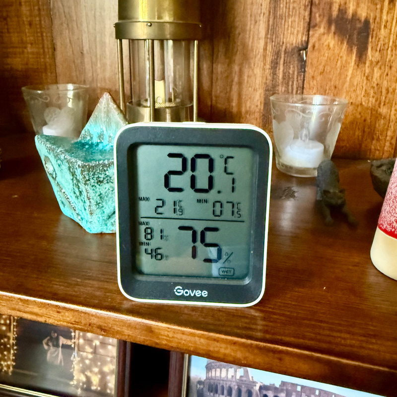 Auto-generated description: A digital thermometer displays temperature and humidity readings on a wooden shelf surrounded by decorative items.