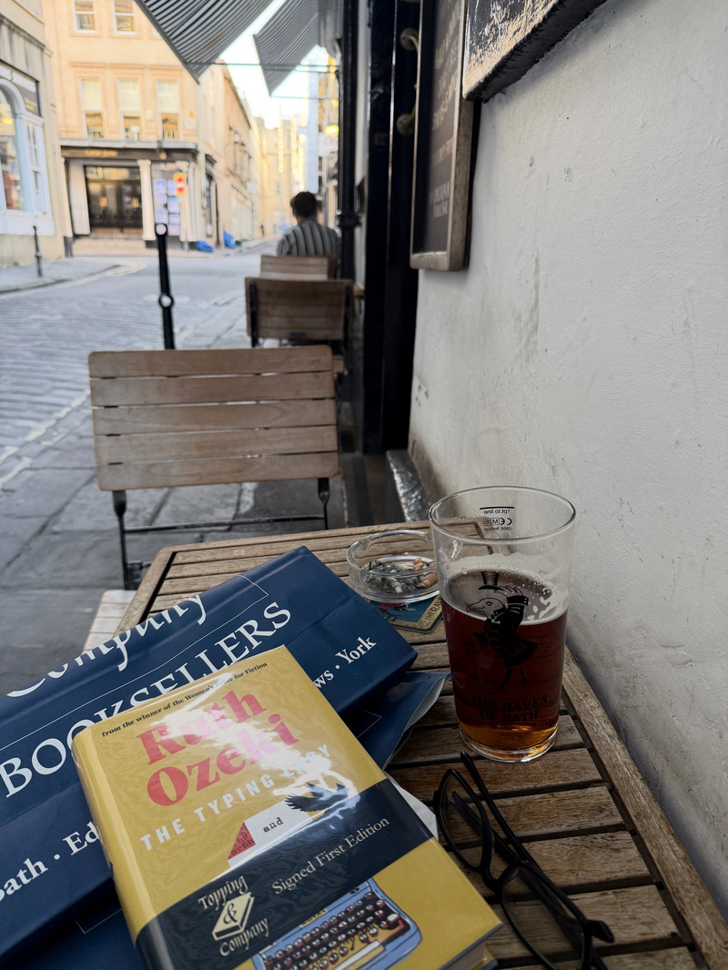 A small outdoor café table holds a book, a glass of beer, an ashtray, and a pair of glasses, with outdoor seating and a street scene visible in the background.
