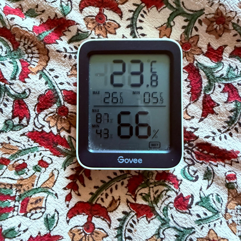 Auto-generated description: A digital thermometer and hygrometer display shows a temperature of 23°C and a humidity of 66% against a floral-patterned background.
