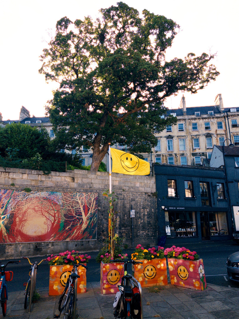 A yellow flag with a smiley face waves above a group of flower pots and a wall painted with smiley faces, set against a backdrop of trees, buildings, and a colorful mural.