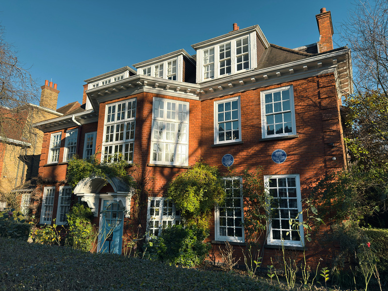 A red-brick house with large windows and a blue door is surrounded by greenery under a clear blue sky.