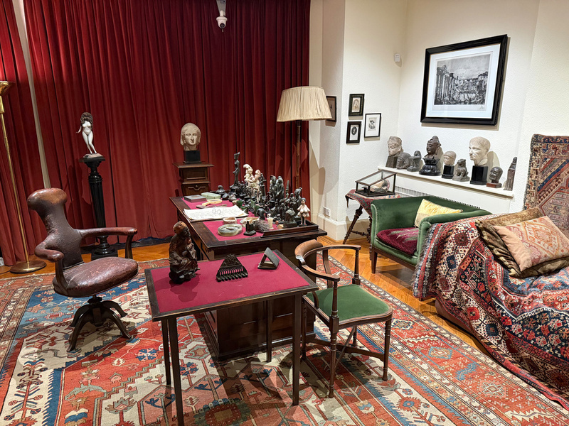 A historical study room is filled with antique furniture, rugs, busts, and art pieces, creating a rich, classical ambiance.
