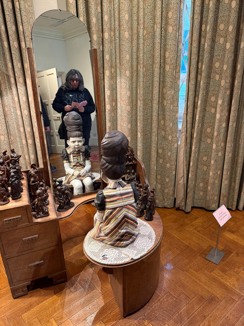 A sculpture of a child sits on a wooden vanity, reflected in the mirror, with decorative figurines surrounding it.