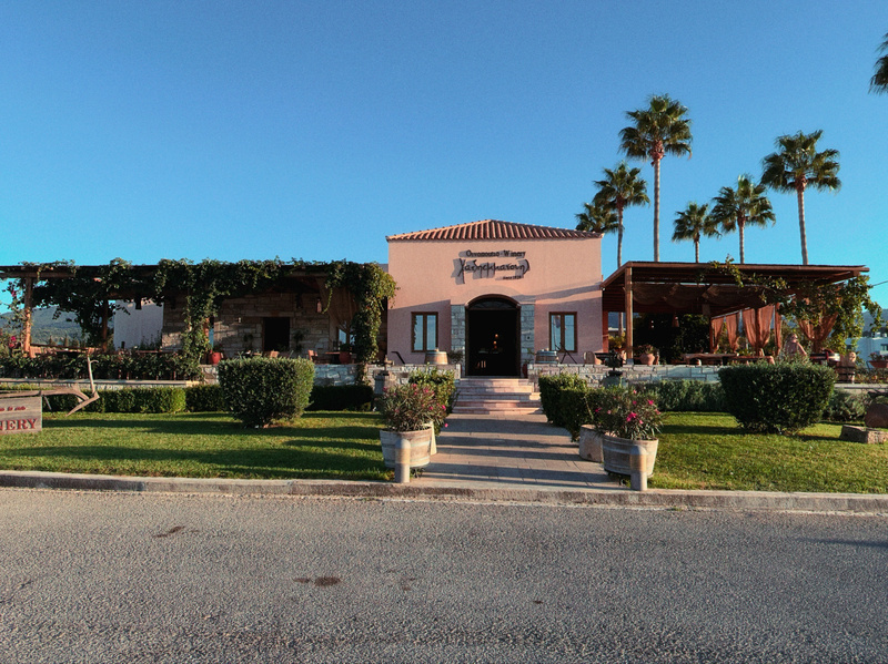 A charming building with a Mediterranean architectural style is surrounded by neatly trimmed bushes, potted plants, and tall palm trees under a clear blue sky.