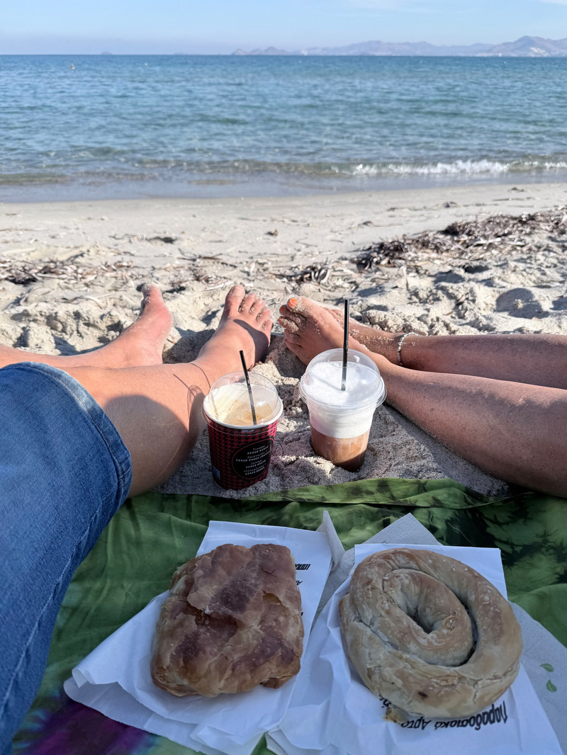 Two people relaxing on a sandy beach with iced coffee drinks and pies laid out on a cloth in front of them, next to the sea.