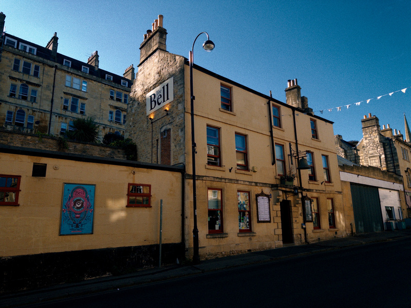 A yellow building with a sign that reads Bell stands along a street under a clear blue sky, with additional older buildings visible in the background.