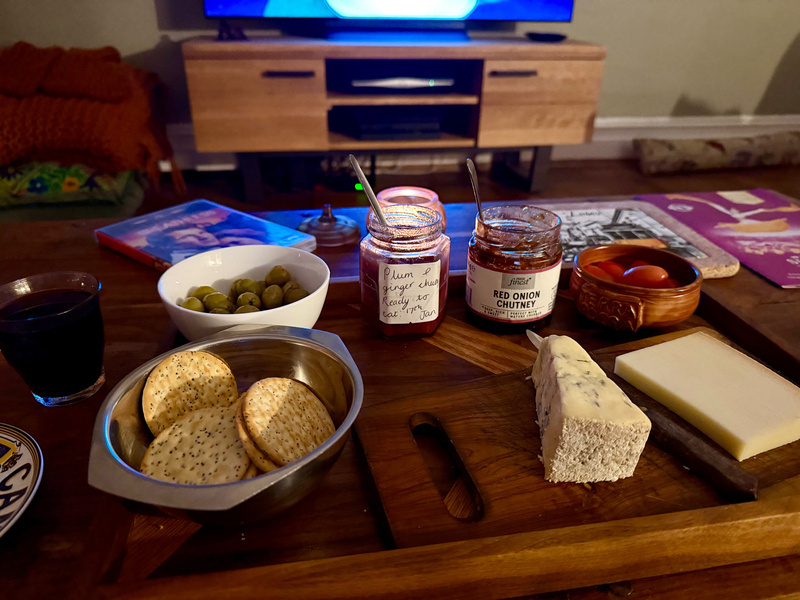 Auto-generated description: A cozy living room setup features a wooden table with cheese, crackers, olives, chutney, and a glass of red wine in front of a television.