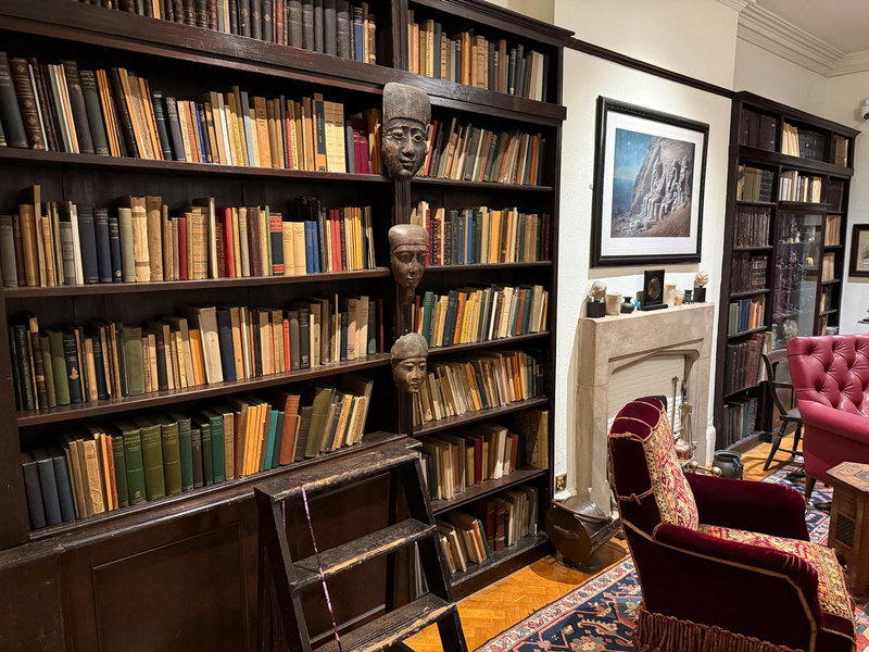 A cozy library features bookshelves filled with books, tribal masks on display, and a comfortable seating area with a red upholstered chair.