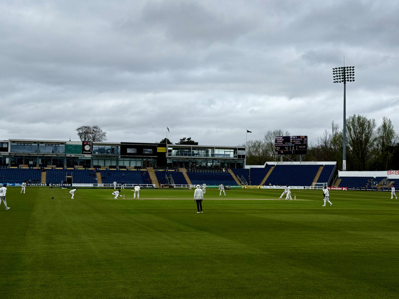Auto-generated description: A cricket match is taking place in a stadium under a cloudy sky with players in white uniforms on a green field.