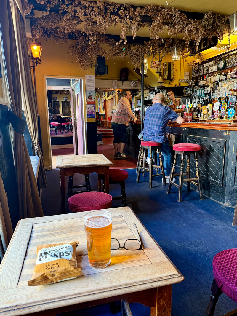 A cozy pub interior features a wooden table with a glass of beer, a packet of crisps, and glasses in the foreground, while patrons chat near the bar in the background beneath hanging dried hops.