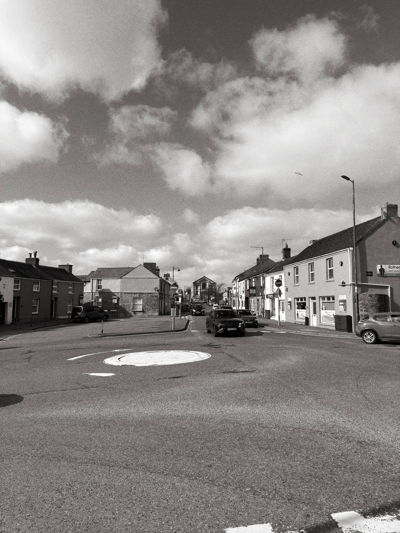 Auto-generated description: A monochrome street scene shows a roundabout in a small town with vehicles, buildings, and cloudy skies overhead.