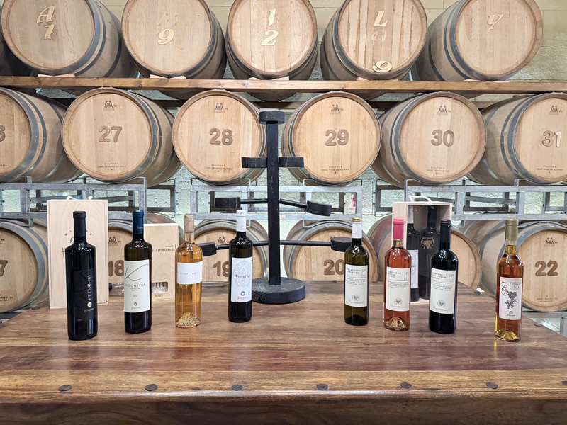 A variety of wine bottles are displayed on a wooden table in front of stacked wine barrels.