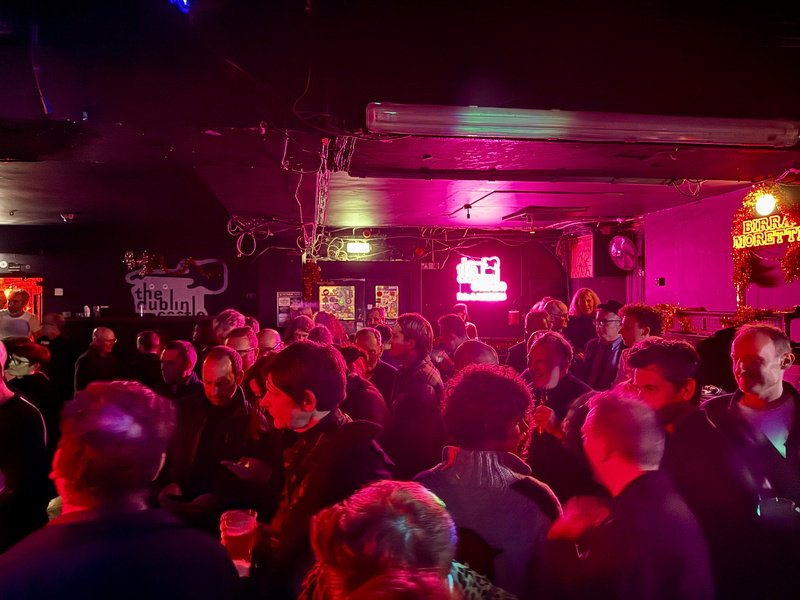 Auto-generated description: A group of people gathers in a dimly lit venue with neon signs and ambient lighting.