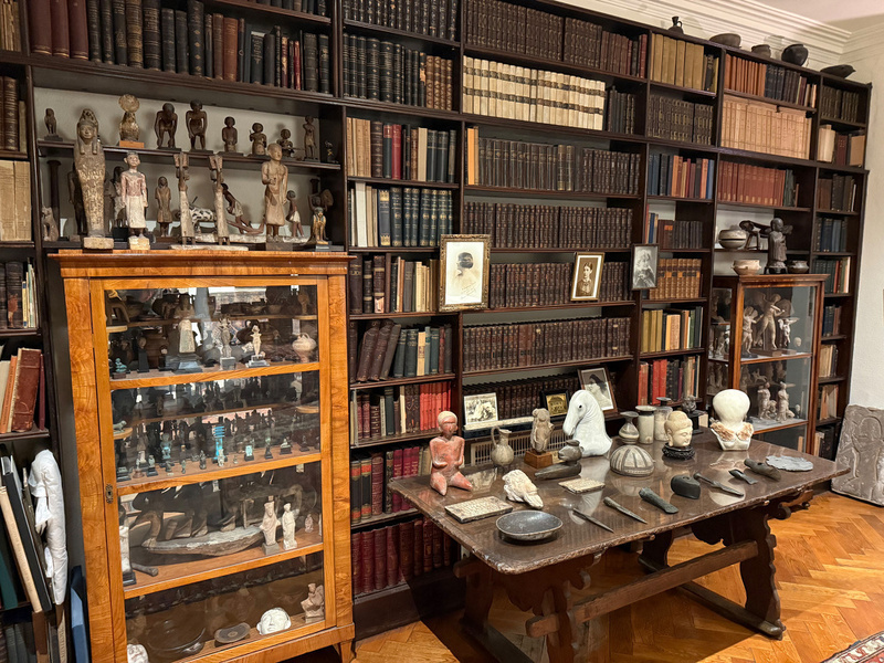 A richly decorated study features bookshelves filled with numerous books and artifacts, alongside a wooden table displaying various sculptures and items.