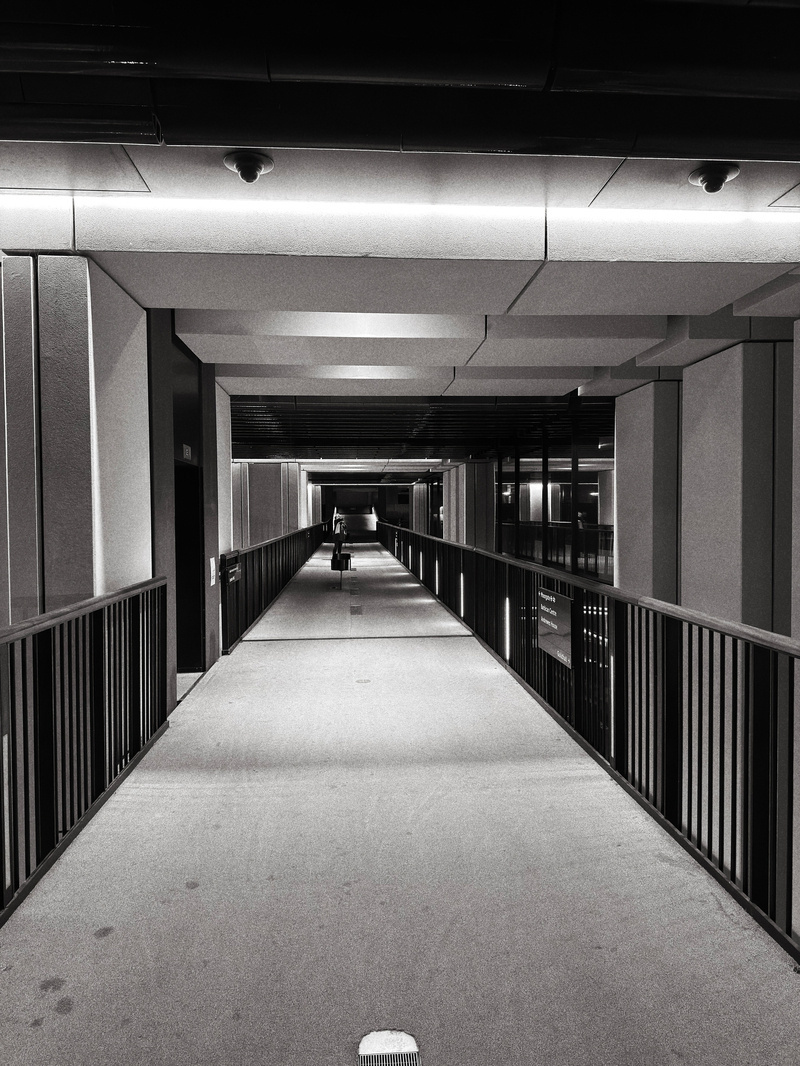 Auto-generated description: A black and white photo depicts a person walking along a modern, well-lit corridor with railings on both sides.