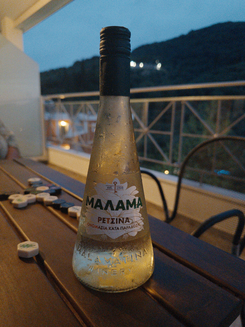 Auto-generated description: A frosted bottle of Malamatina Retsina wine is placed on a wooden table with game pieces scattered around, set against an outdoor evening backdrop with a railing and distant hills.