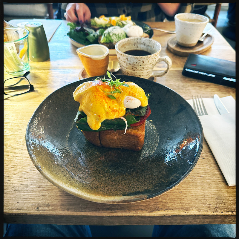 A delicious eggs Benedict dish with hollandaise sauce is served on toast, placed on a rustic plate, accompanied by various cups and utensils on a wooden table.