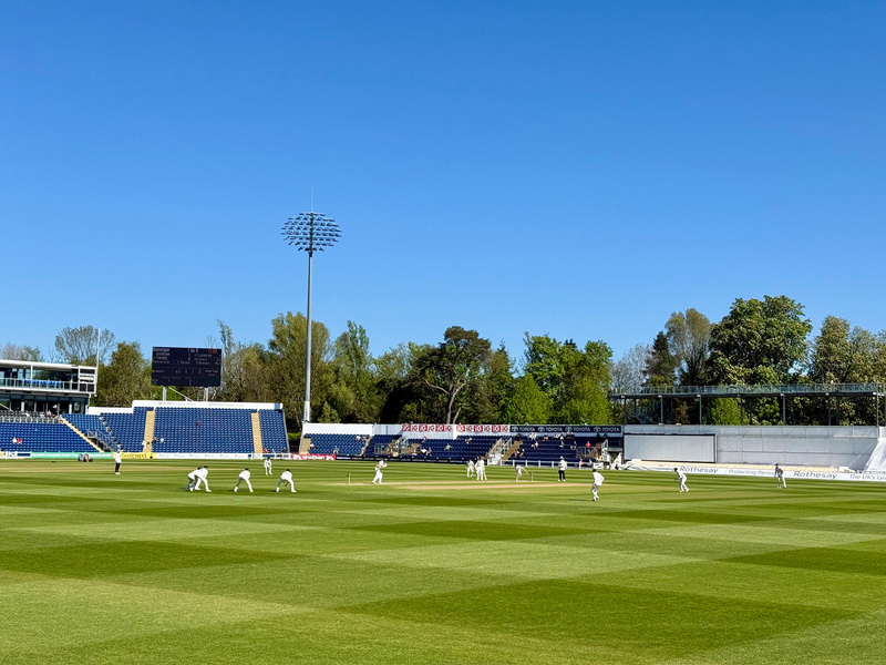 A cricket match is taking place on a sunny day in a stadium surrounded by trees.