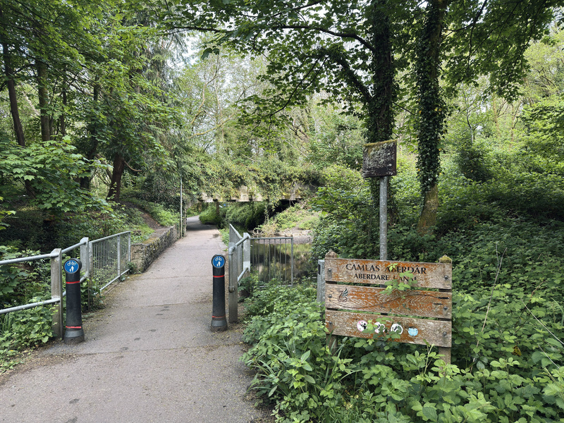 Footpath leading under a bridge covered in foliage. Sign titled Aberdare Canal.