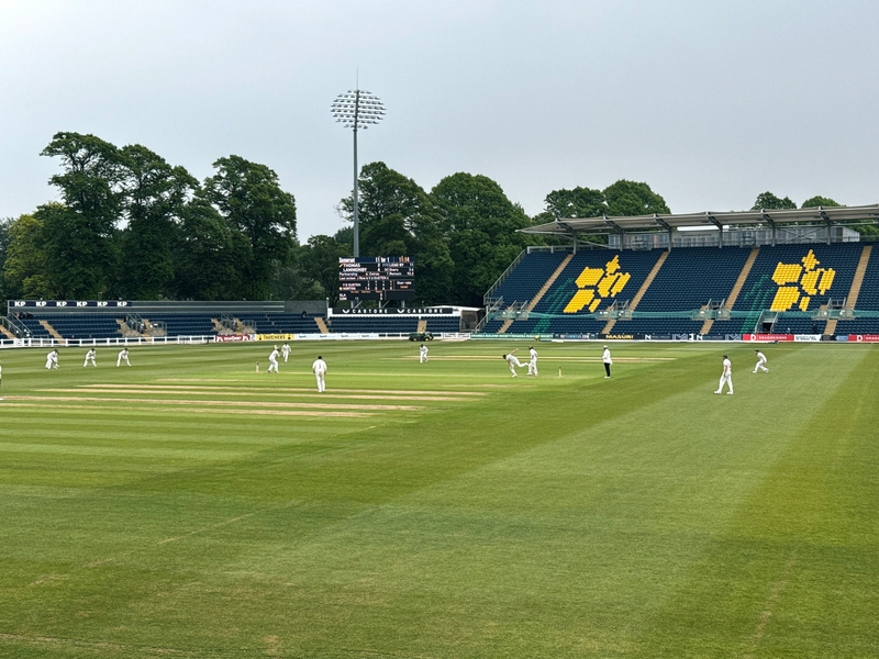 A cricket match is taking place in a stadium with players on a grassy field, surrounded by empty stands and large trees in the background.