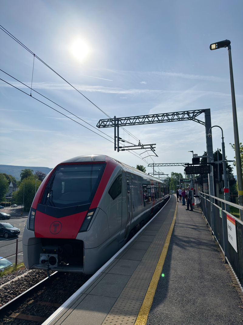 A sleek, modern train is parked at a sunlit station platform with a few people standing nearby.