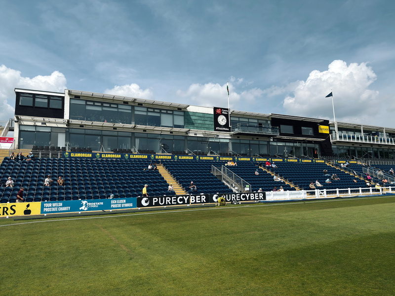 A sports stadium features a seating area in front of a modern building, with a lush green field in the foreground and a cloudy sky above.