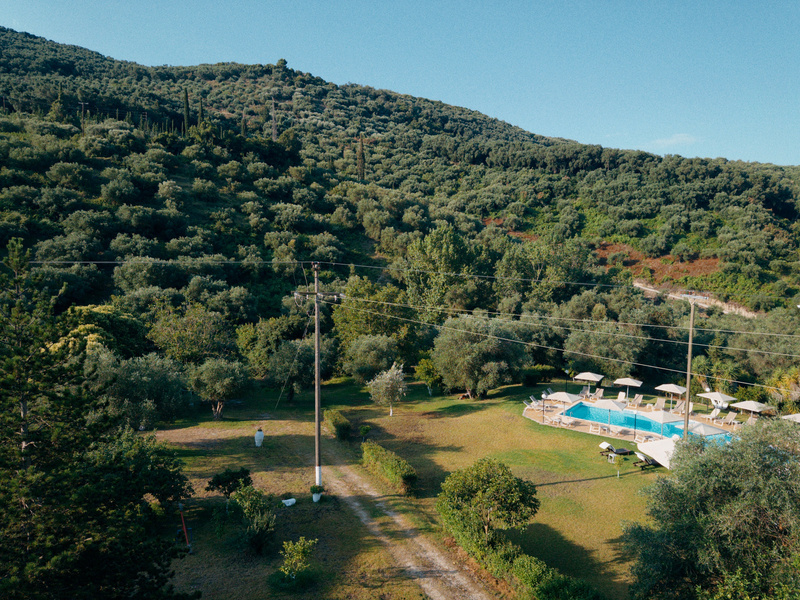 A scenic hillside covered with dense trees surrounds a grassy area featuring a swimming pool with umbrellas and lounge chairs nearby.