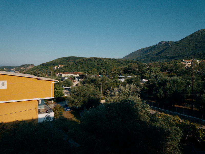 A peaceful rural landscape features green hills, scattered houses, and a bright yellow building under a clear blue sky.