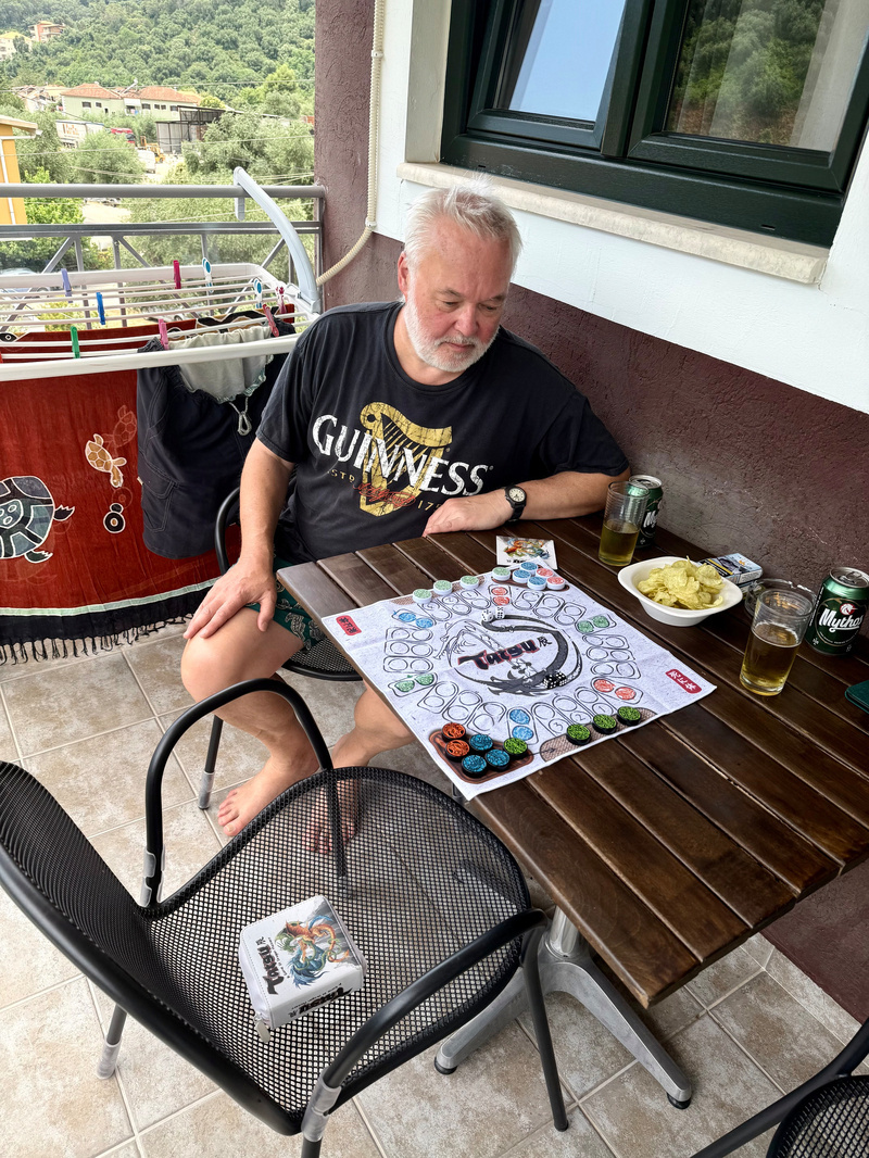 A man wearing a Guinness t-shirt is sitting at a table on a balcony playing a board game named Tatsu, with snacks and drinks beside him.