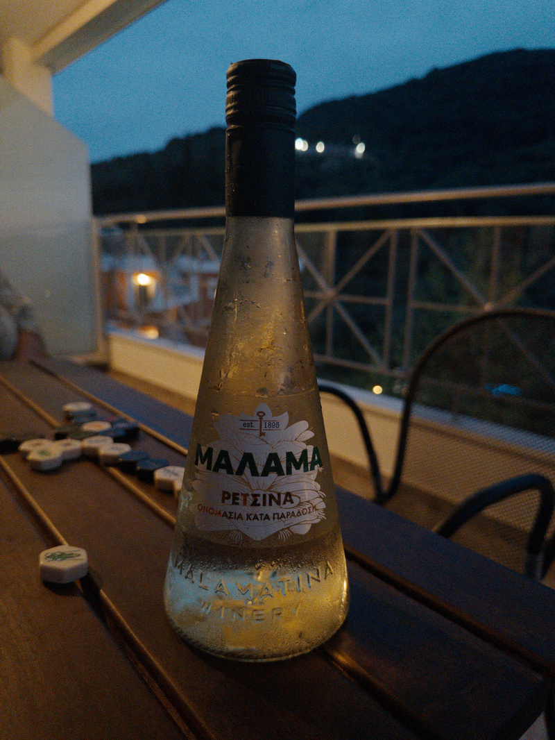 A frosted bottle of Malamatina Retsina wine is placed on a wooden table outdoors, with a scenic evening background.