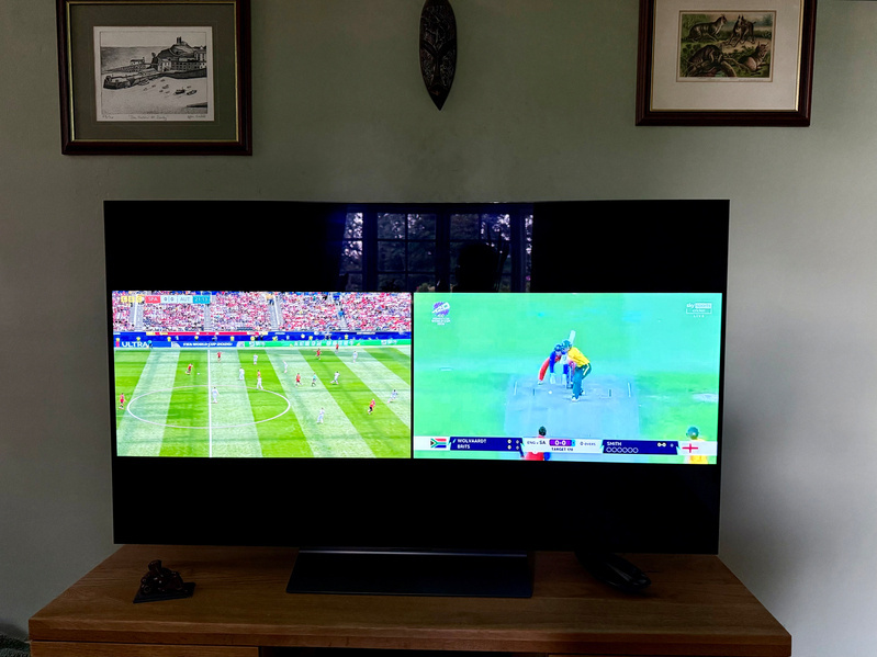 A television on a wooden stand is displaying a split screen with a soccer match on the left and a cricket match on the right.