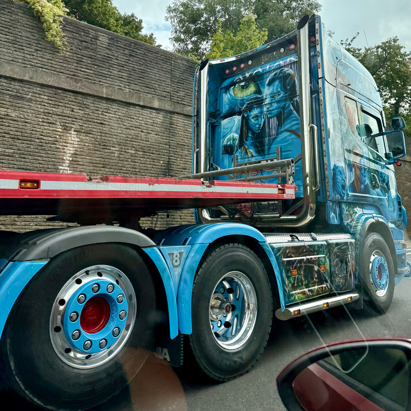 A blue semi-truck features a detailed artistic design with an Avatar motif on the cab, and it is passing by a stone wall with greenery visible above.