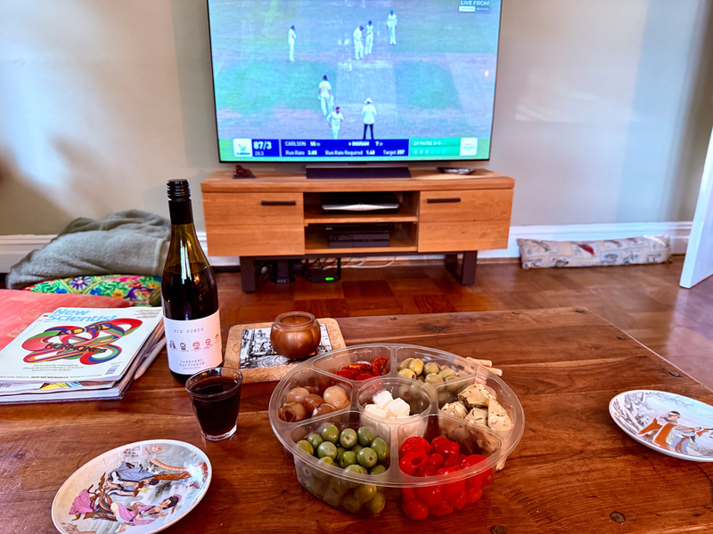 A cozy setup with a glass of red wine, assorted snacks on a platter, and a cricket match playing on the television in the background.