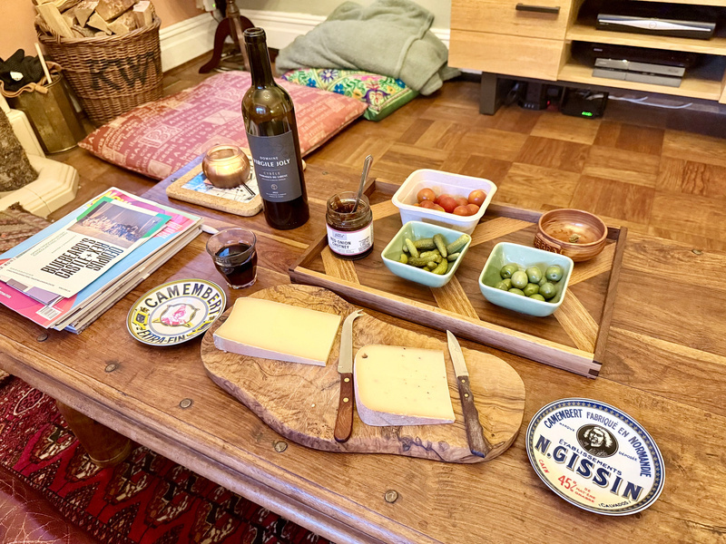 A wooden table is set with various cheeses, pickles, olives, cherry tomatoes, a bottle of red wine, a glass of wine, jars of condiments, and some reading materials in a cozy living room.