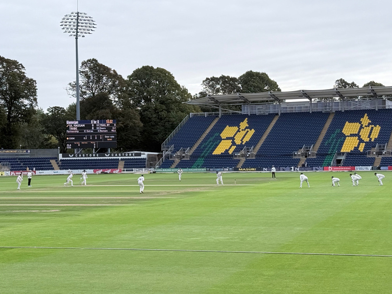 A cricket match is in progress at a stadium with players positioned on the field and spectators' seating visible in the background.