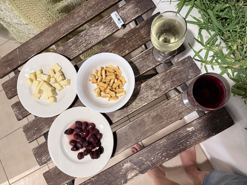 On a rustic wooden table, there are three white plates containing cheese cubes, small fish-shaped biscuits, and black olives, accompanied by a glass of white wine and a glass of red wine.
