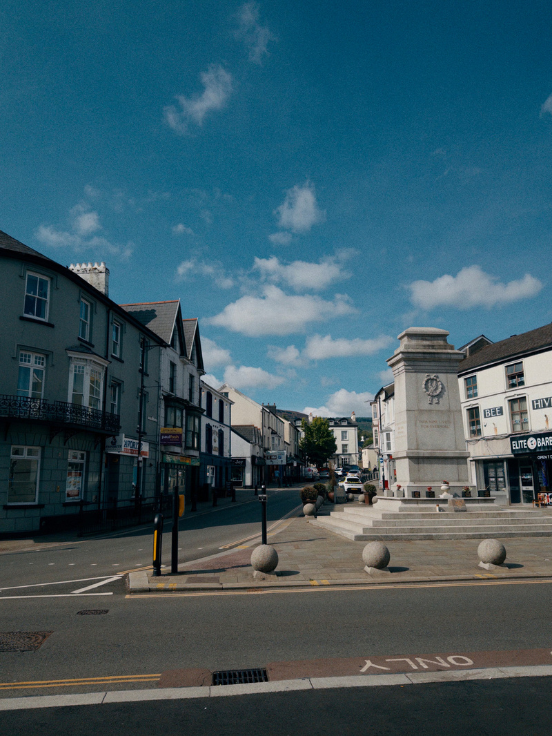 Auto-generated description: A quiet town street with shops on either side and a stone monument in the center under a blue sky with scattered clouds.