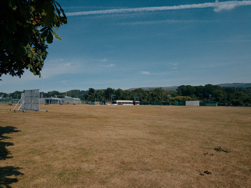 Auto-generated description: A large, open grassy field with some trees, sports equipment, and buildings in the distance under a blue sky.