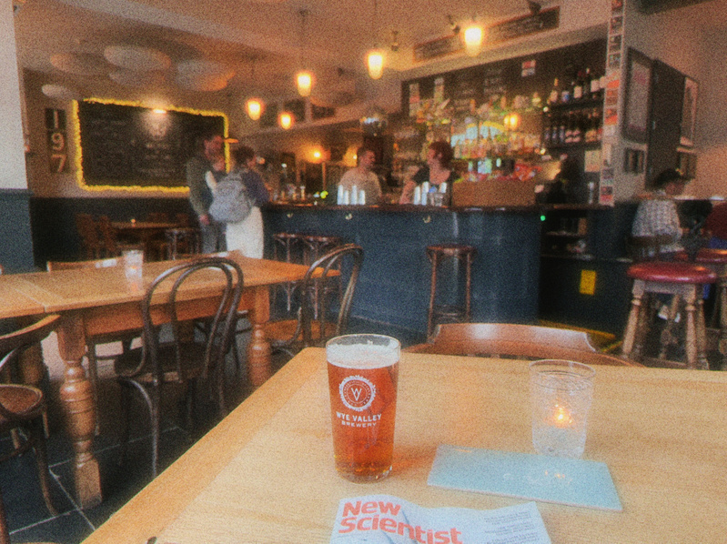 A pint of beer from Wye Valley Brewery, a glass of water, and a copy of New Scientist magazine are on a wooden table in a cozy pub with people chatting at the bar in the background.