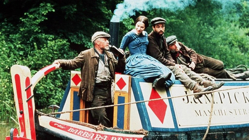 A group of people dressed in period clothing are riding on a colorful narrowboat traveling through a wooded area.